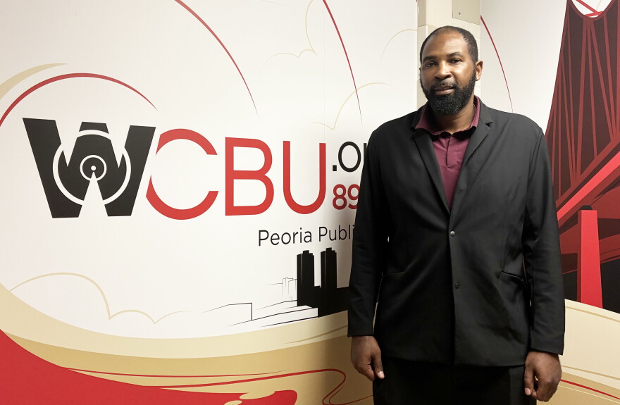 A man in a black blazer and maroon shirt stands in front of a WCBU 89.9 Peoria Public Radio wall with abstract city and bridge artwork.