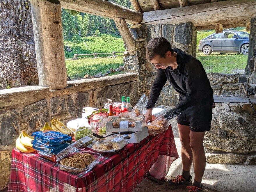 A hiker stands at a table full of various foods inside a covered shelter. The hiker is pulling a piece of bread out of a bag.