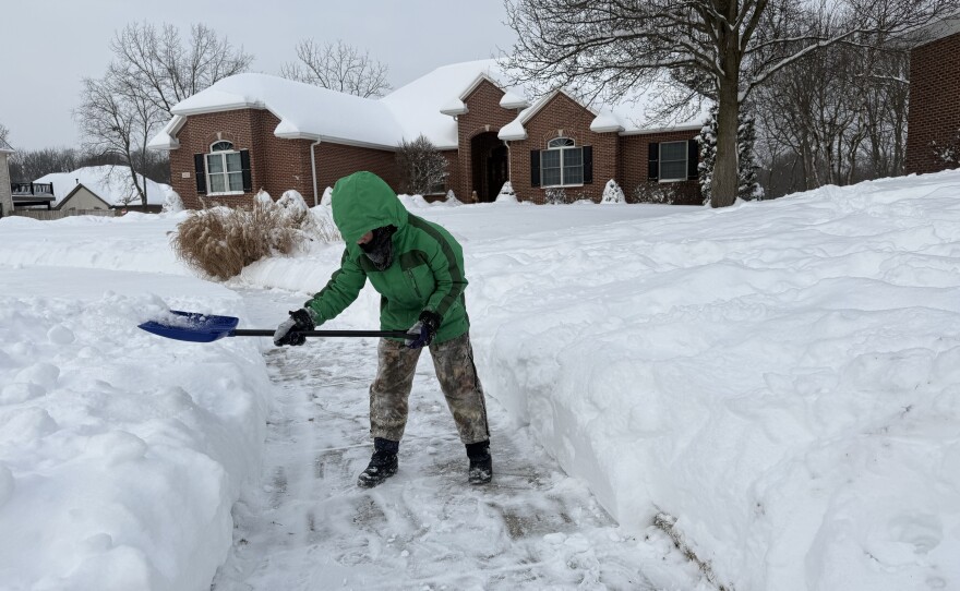 A child is surrounded by snow. He's wearing a bright green winter coat, camo snow pants, boots and gloves is shoveling snow off the sidewalk.