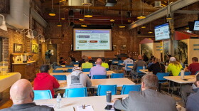 Water utility workers sit at long tables lined up in rows inside a century-old, brick-walled water-pumping station that has been converted into an event space. They look at a large screen and a person speaking at a podium.