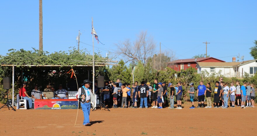 Photo of a man wearing jeans and a decorated vest stands in the middle of a corral with red dirt holding a flag. A group of young boys stands behind him.