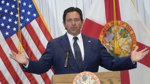 A man in a suit speaks at a podium during a press conference with arms spread wide