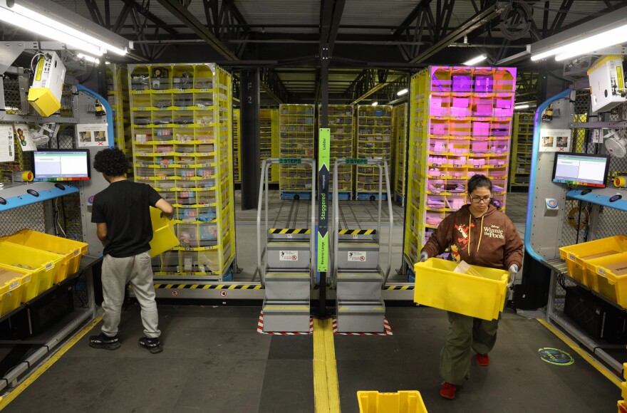 Amazon associates are shown packing inventory into tall yellow pods with multiple small cubicles. One of the pod is illuminated in pink, and the associate is walking away with an empty tote.