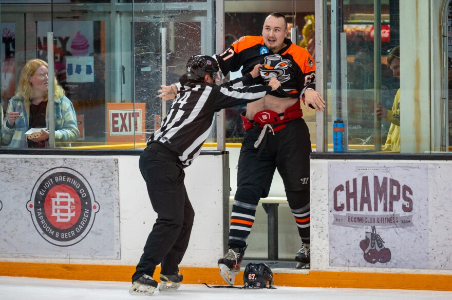 Linesman Kyle Cedrone pushes Danbury Hat Tricks forward Jesse Swanson into the penalty box after the second fight during the first period of the Hat Tricks’ home opener on October 10, 2025. Swanson was part of both first-period fights and received four penalties, including a 10-minute misconduct penalty. The first fight occurred just two seconds into the game.(Jonathan McNicol/Connecticut Public)