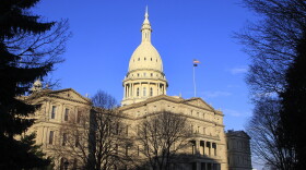 The dome of the Capitol glows in bright natural light, against a deep blue sky with bare trees on the grounds