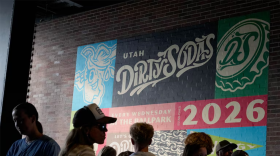 A colorful mural on a brick wall reads "Utah Dirty Sodas" and "2026." A crowd of people stand in front of it, facing different directions. 