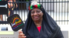 An older Black woman wears a headscarf and a festive crown that reads "Juneteenth" She also holds a paddle fan that reads "Juneteenth, commemorate, educate, celebrate." 