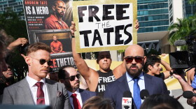 Two suited men speak on microphones while men behind them hold up signs. 