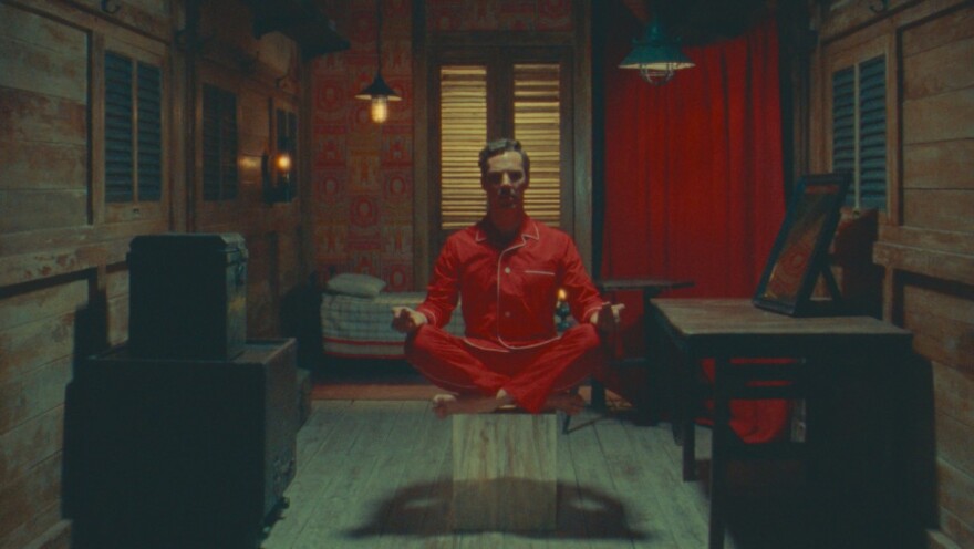 A man sitting cross-legged in his pajamas appears to float above the floor in a small room with wooden walls and a wooden floor.