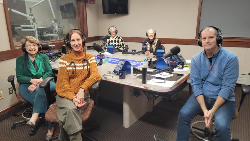 Five people wearing headphones sit at a table in a radio talk studio: a woman front left has short brown hair and is wearing glasses, a green sweater, green scarf, green pants and brown shoes: a woman front right has shoulder-length brown hair and is wearing an orange sweater with two white stripes across the chest and arms and two black ribbon bows on the chest, brown pants and brown boots; a woman back left has short grey hair and is wearing glasses and a sweater with a black-and-white diamond pattern over a white button-down shirt; a woman back right has short grey hair and is wearing a gold puffy vest over a black turtleneck sweater; a man at center has very short dark hair and is wearing a light bluee quilted sweatshirt and blue jeans.