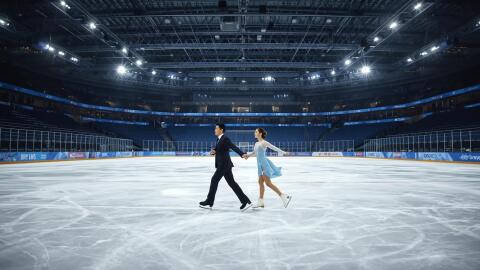 Skaters, a man in a tuxedo-style outfit and a woman in a flowing blue costume with white sleeves take to the ice