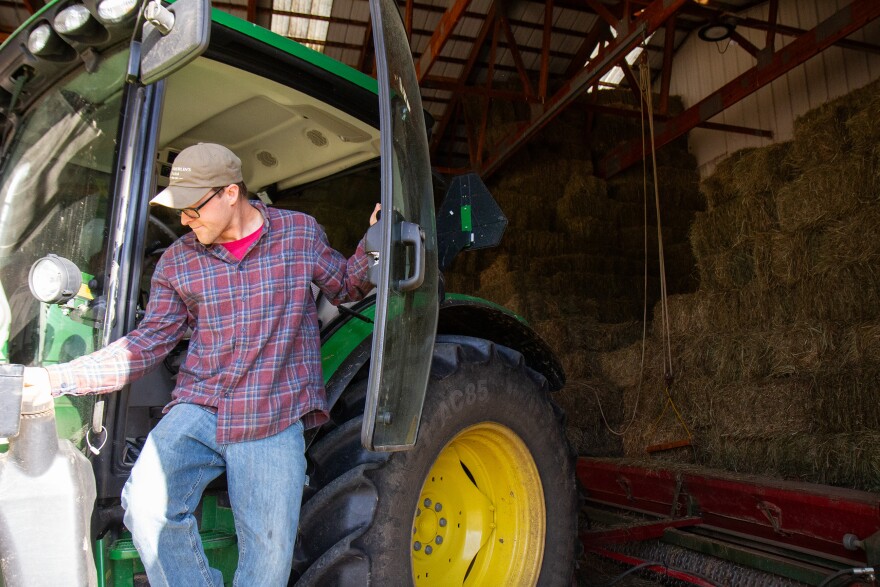 A photo of a person wearing a baseball hat, glasses, plaid shirt and jeans steps down from a big green and yellow tractor inside a barn.