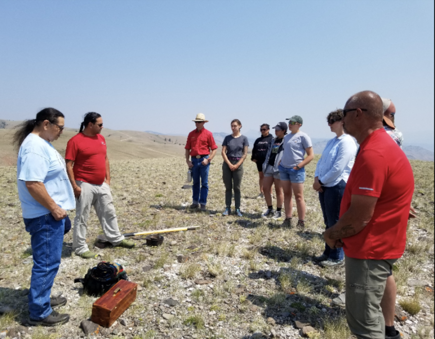 A group of people stand in a circle around a backpack, toolbox, and shovel. They're on a high rocky hill with blue skies around them.