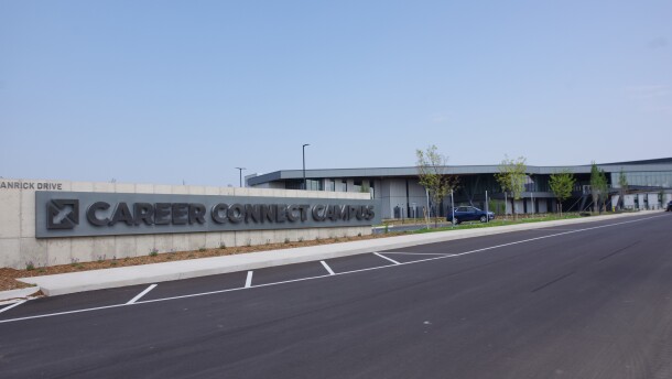 A large, shiny gray sign reads "Career Connect Campus" in black text. A large and long building looms behind the sign, with rows of glass windows and pale gray metal siding lining the outside of the facility. 