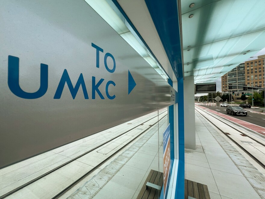 A chrome sign is seen in foreground with blue lettering that reads "To UMKC." The sign sits under a covered space that looks like a stop for mass transportation. Streetcar tracks can be seen on either side of the sign.
