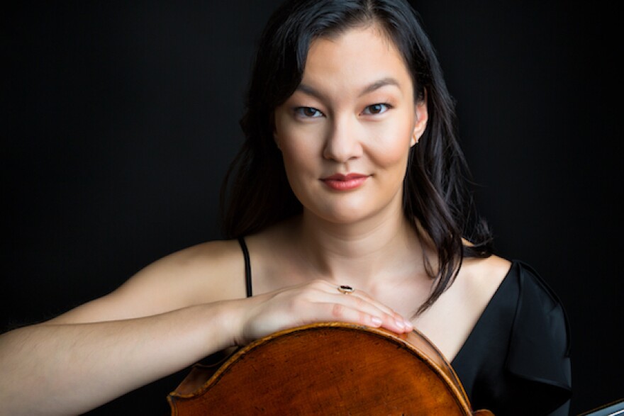 Cellist Shirley Hunt is seating looking straight into the camera, wearing a black dress, with her cello across her lap and her left hand wresting on its curve