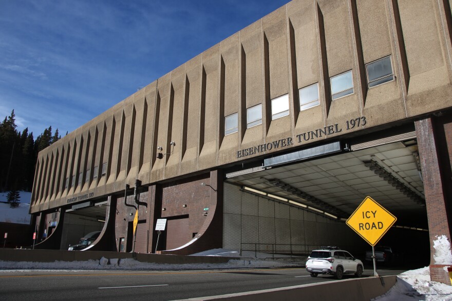 A brown tunnel has a roadway underneath it with a white car driving through. There's a sign that says "ICY ROAD." Over the top of the tunnel reads "Eisenhower Tunnel 1973."