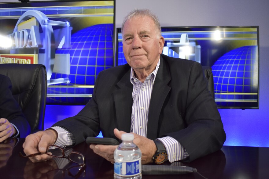 Clark is wearing a suit and sitting at a table. He’s posing for a photo and holding his glasses in his hands.