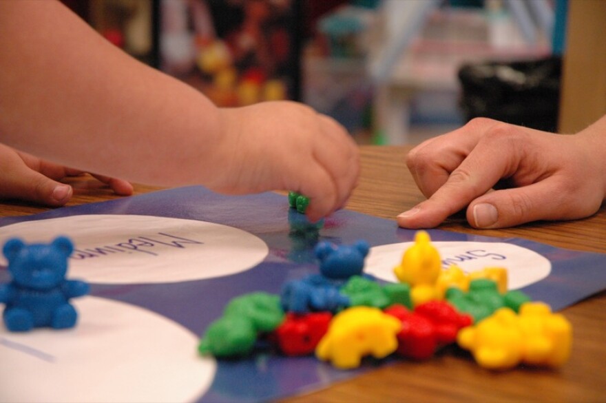 A child's hand reaches to a pile of red, yellow, green and blue bears and moves them across a sheet of paper toward an adult's hand 