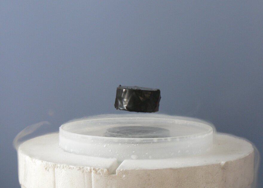 A black magnet levitates slightly above a clear dish which sits on top of a block of liquid nitrogen. 