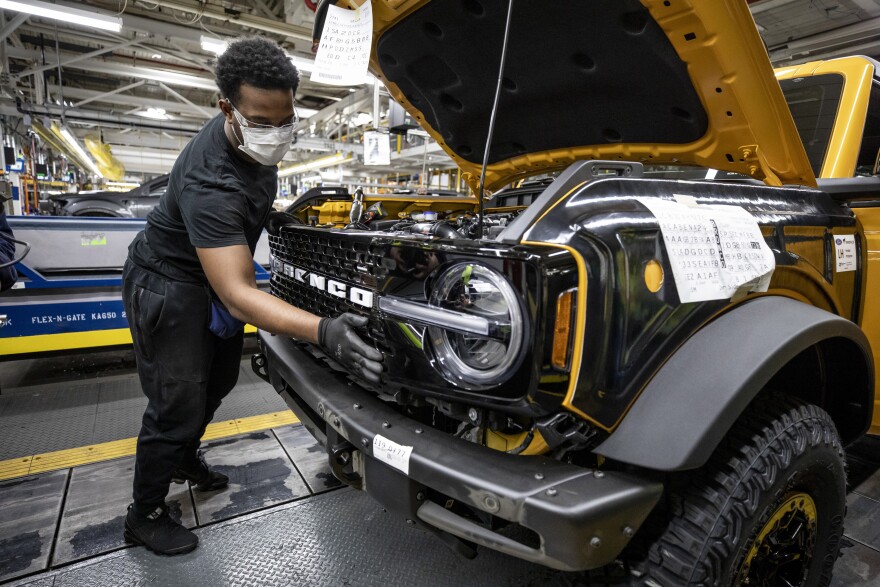 Ford employee works on the front of a new Ford Bronco at an assembly plant. He wears a mask.