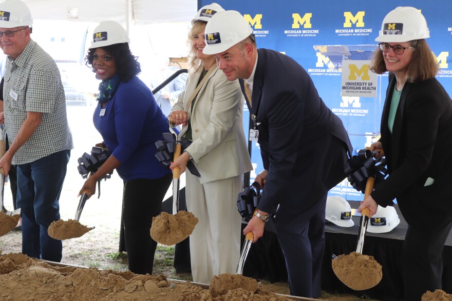 A few people in business attire wear hard hats and hold shovels, smiling at a camera and digging up dirt.