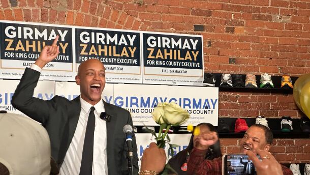 A man standing on a podium in front of a crowd points at the ceiling with his right hand while talking and smiling.