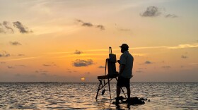 A man stands in the ocean, in shadow after sunset, with an easel in front of him. 