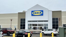 Large tan box building with the bright blue and yellow IKEA brand logo on the front, people walking to and from the glass entrance doors into a large parking lot with several cars and yellow posts in foreground.