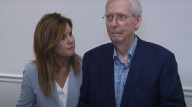 a woman stands next to senator mcconnell as he seems to stare off in the distance