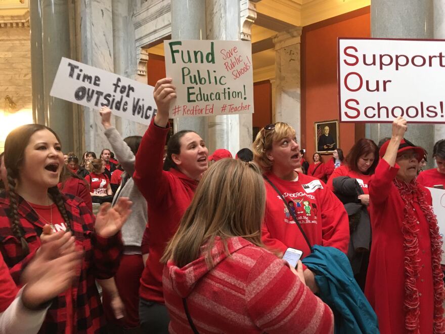 A crowd of adults wearing red and cheering inside a marbled building. Some are holding handmade signs that say "time for outside voices," "fund public education," and "support our schools."