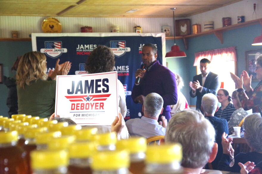 John James, a man wearing a purple quarter-zip shirt, stands among a crowd of people who are clapping -- some are on their feet, and some are waving signs that say "James + Deboyer for Michigan". He is in a room with farmhouse-themed decorations, and there is a large banner behind him that reads "John James for Governor, Michigan". 