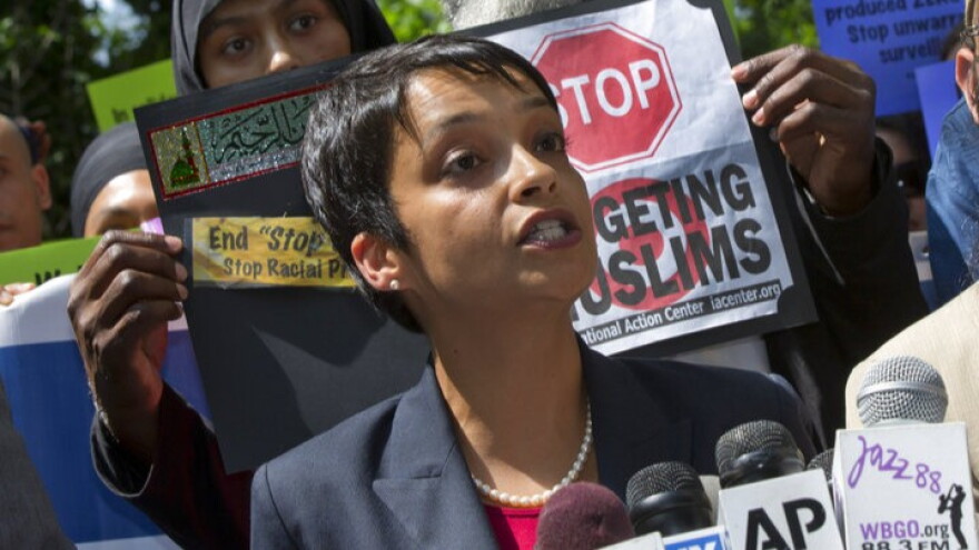 A young woman with short dark hair and dark complexion in a gray blazer and pearl like necklace speaking behind microphones with people behind her holding up signs about targeting muslims. 