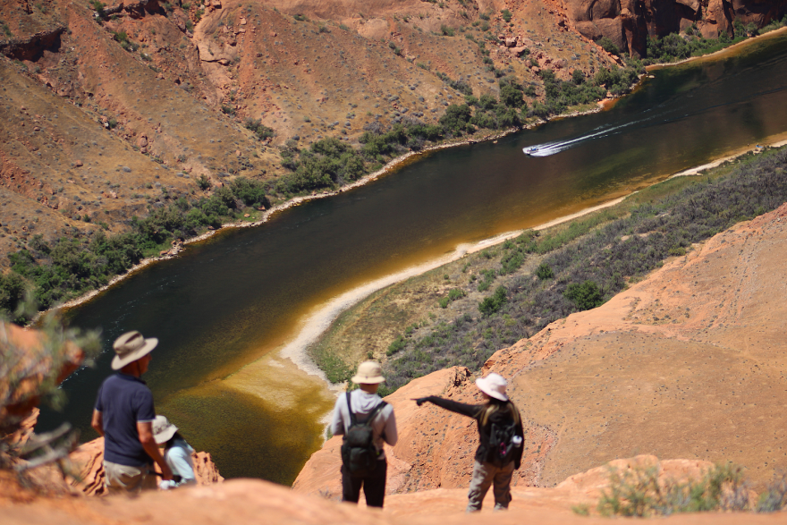 Tourists stand on a tall rocky outcropping, looking down at a river as it starts to bend around the outcropping. There's some greenery right at the banks, but the landscape is otherwise red and dry.