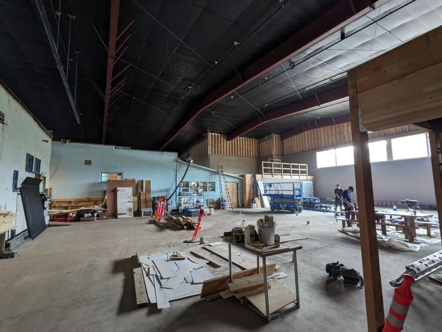 A construction site, with materials and tools strew about. There's a large metal roof arching over and encasing a whole other building to the left.