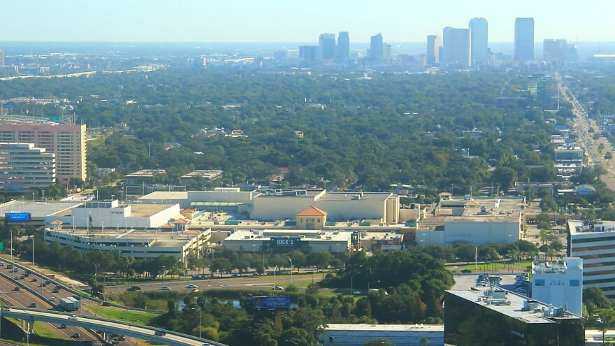 an aerial image showing a large interior shopping mall in the foregrown and downtown skyline through hazy sky toward the rear of the frame.  A major road on the right of the mall and image frame 