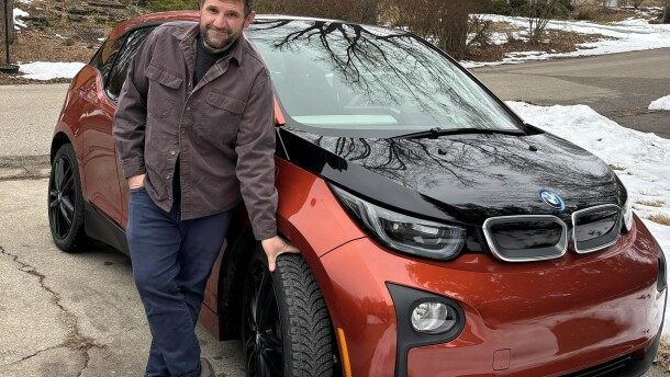 Tyler Lake in a brown chore shirt and jeans standing next to a reddish-brown and black BMW car in a concrete driveway. One hand is in his pocket, one is grabbing a front tire. View of a suburban neighborhood with lingering snow is visible in the background. 