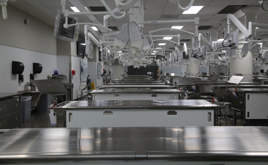 A photo of the dissection lab. Rows of silver tables with lamps, stools, monitors, and chart stands.