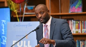 Man wearing a gray suit gestures with his left hand while talking at a podium in front of shelves filled with books and a sign that reads "Full Accreditation."