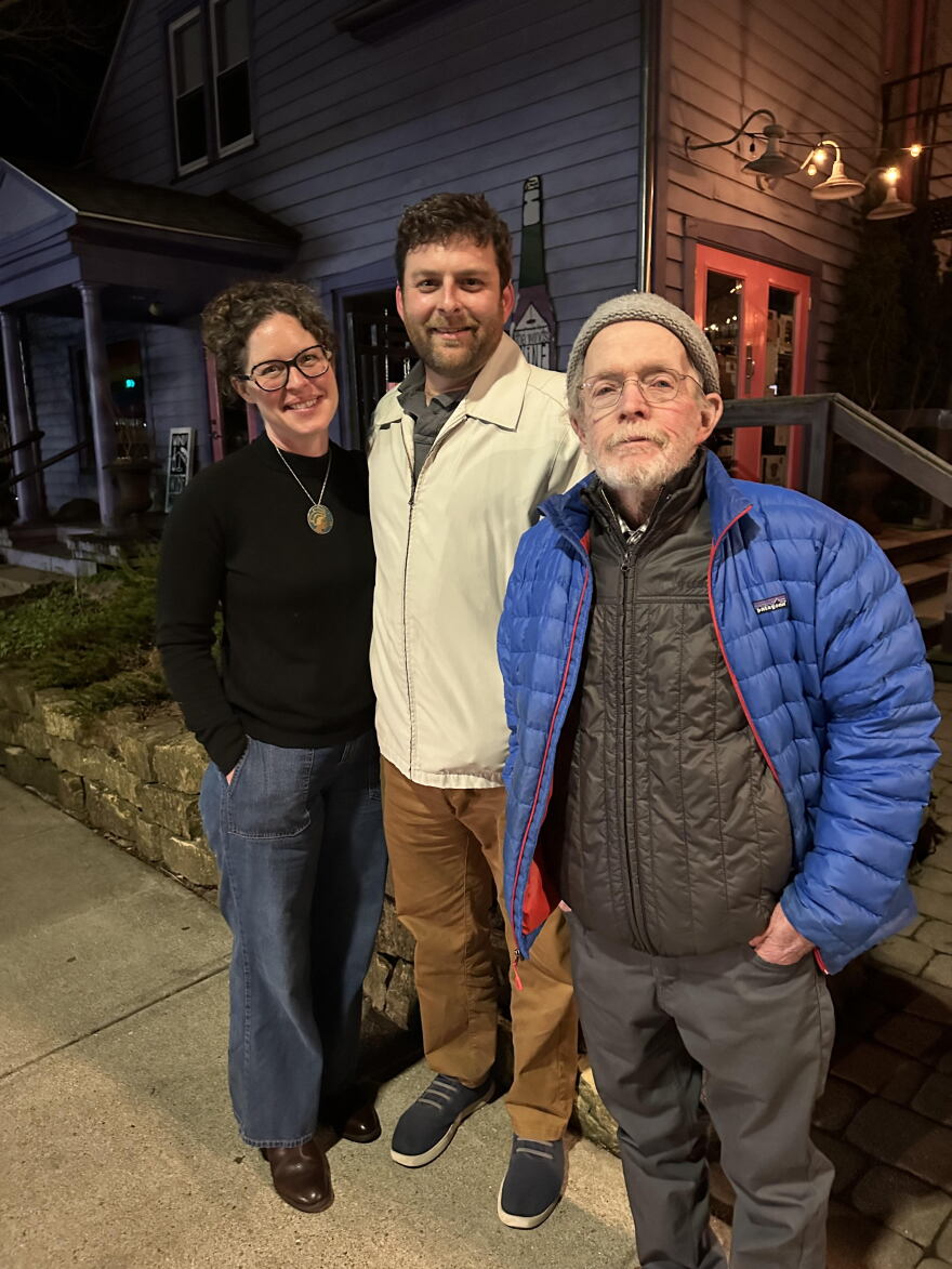 Three people stand on a sidewalk in the evening.