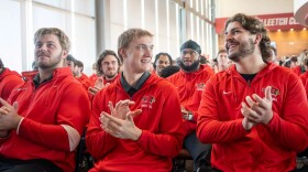 Members of a college football team applauding while wearing red hoodies and seated in a conference room