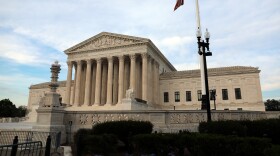 A large, faded white neoclassic style building looms in the center of the image. Several tall pillars hold up the center of the building. A red, white and blue American flag can also be seen flying in the foreground, to the right of the image. 