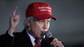 Three-quarter head-and-shoulders view of a man with white hair wearing a dark suit with white shirt, red tie and red "Make America Great Again" hat, as he speaks into a microphone while gesturing with his right index finger raised