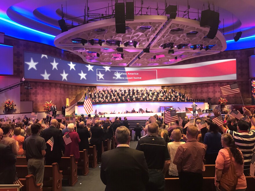 A large room with a big American flag waving on a screen is full of people waving their own American flags.