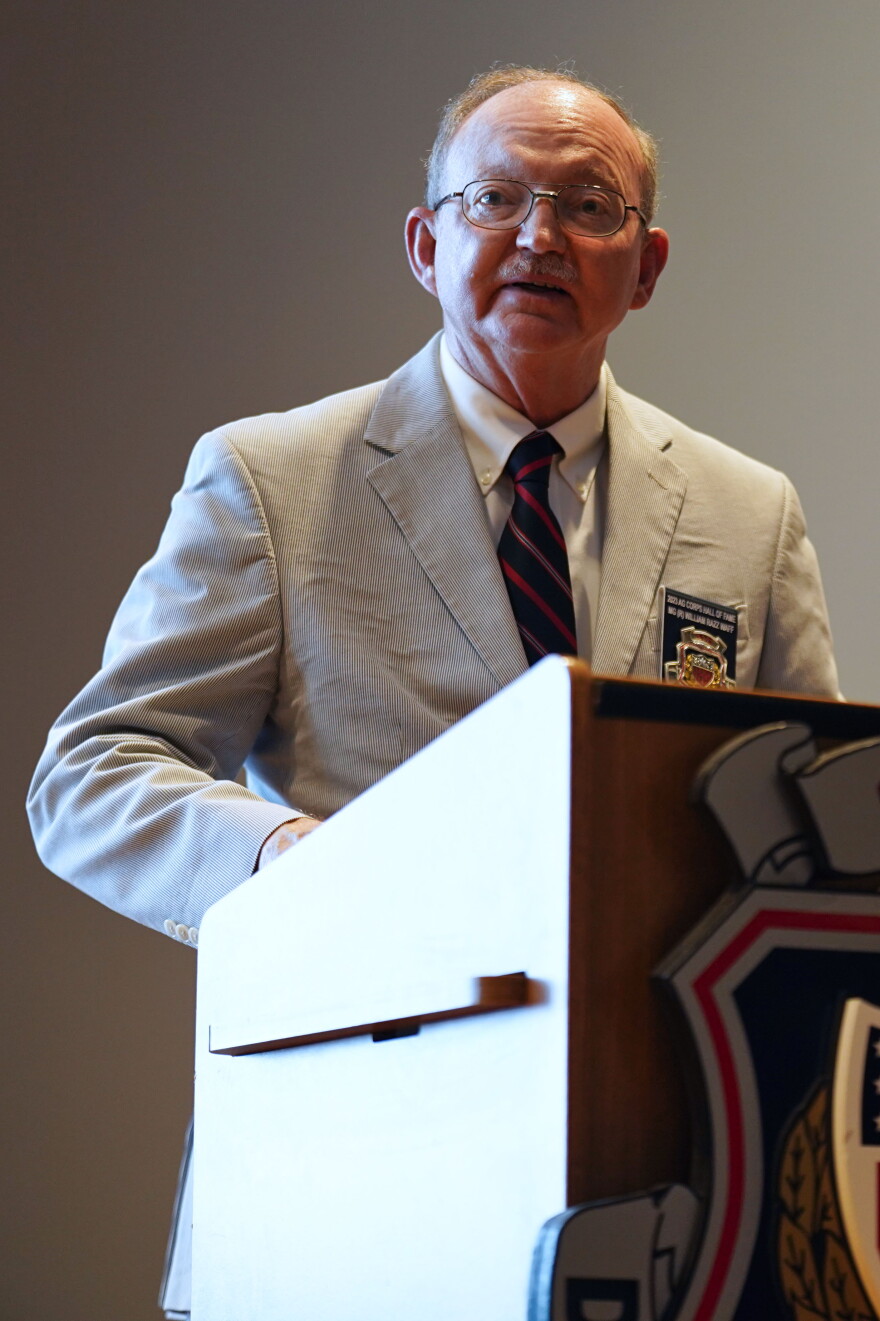 A man in a suit and tie stands behind a podium speaking. 