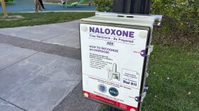A white, black and red box containing free doses of naloxone, a nasal spray medication to reverse drug overdoses, is installed at on a metal pool in a park. A concrete walking path sits next to it on the left, with childeren at play in the background of the top right. On the other side of the box, a patch of grass, leads to a small bush. 