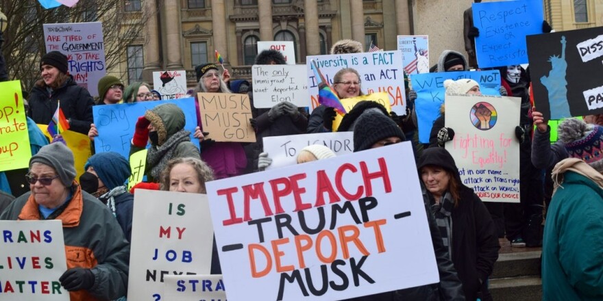 A diverse group of protesters stand outdoors holding colorful signs with messages such as “IMPEACH TRUMP DEPORT MUSK,” “Diversity is a FACT,” and “Trans Lives Matter,” in front of a government building, rallying against tariffs and impacts on economic activity.