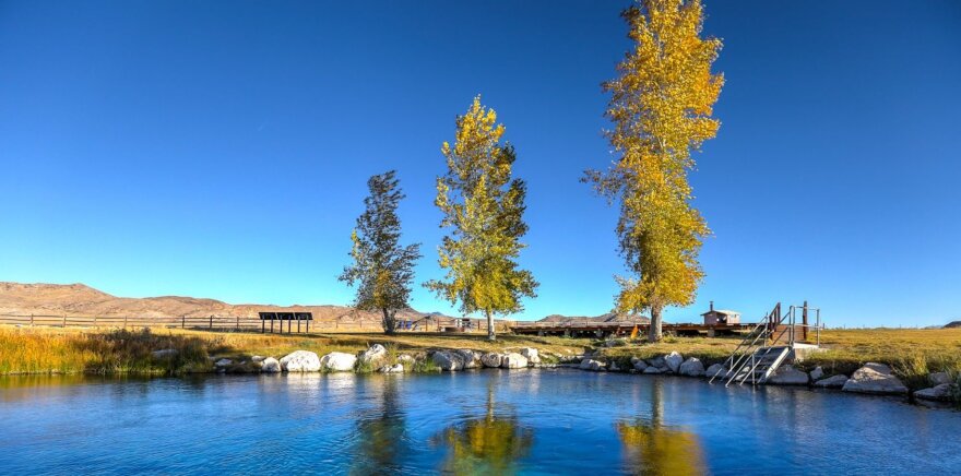 The image shows a hot spring shaded by a few trees on the Duckwater Shoshone reservation in Nye County, Nev. Clear blue skies and a mountain range are in the background.