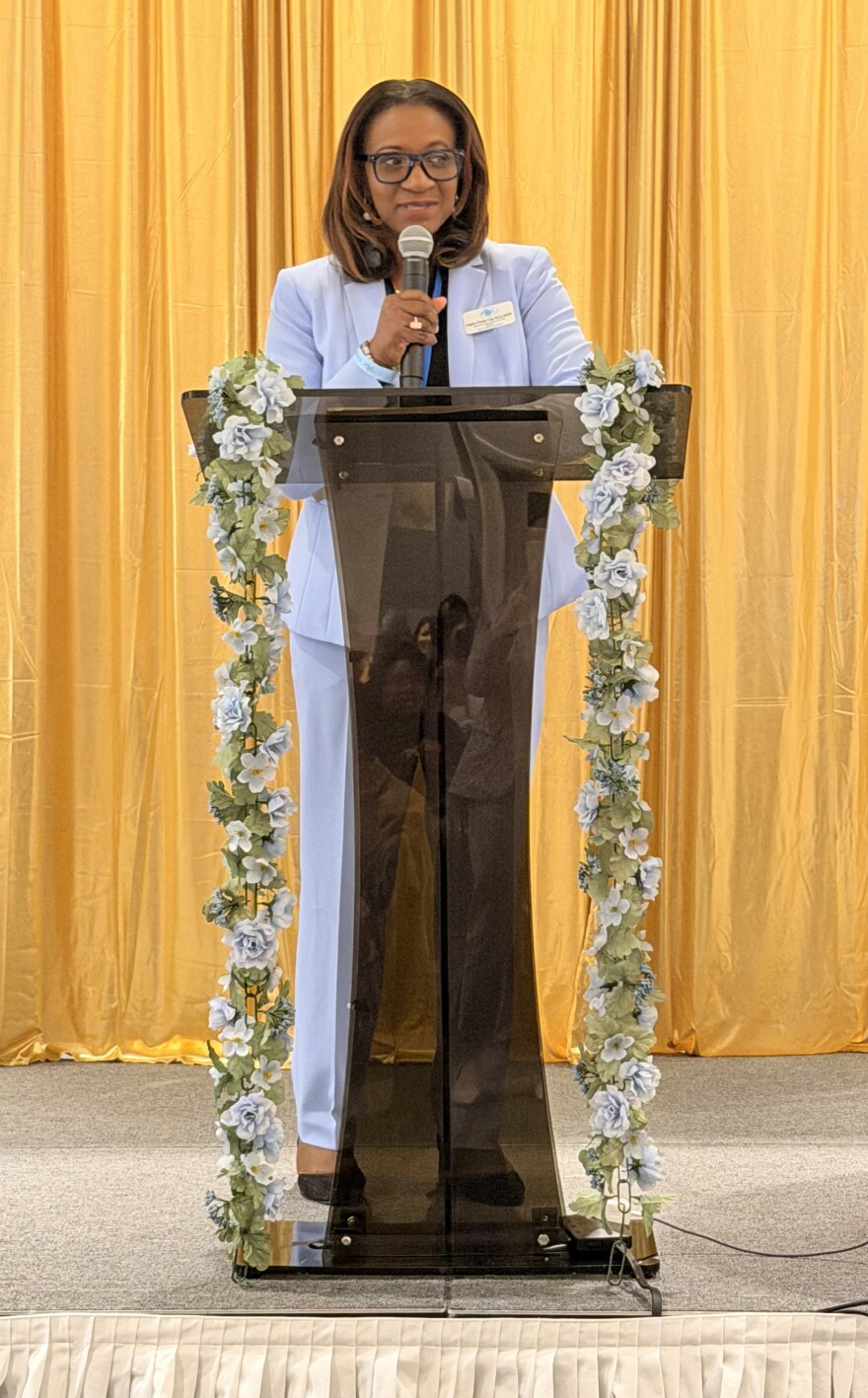 A woman in a light blue suit speaks into a microphone at a podium decorated with blue flowers.
