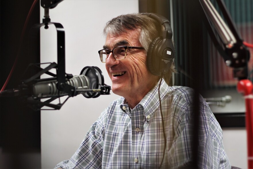 A man wearing glasses and a plaid shirt smiles while seated behind a microphone for a radio interview.